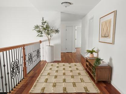 Beige flat-weave area rug with olive-green rectangular stripes along a hardwood hallway beside a wooden console and potted plants.