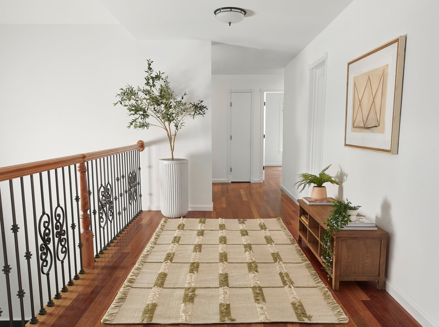 Hallway with warm wood floor, beige striped runner, iron railing, tall potted plant, and wooden console with books.