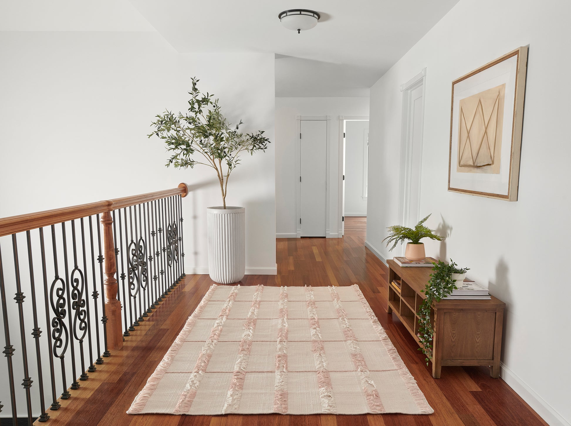 Bright hallway with wood floor, pink-beige striped rug, black iron railing with wooden handrail, potted plant, wall art.