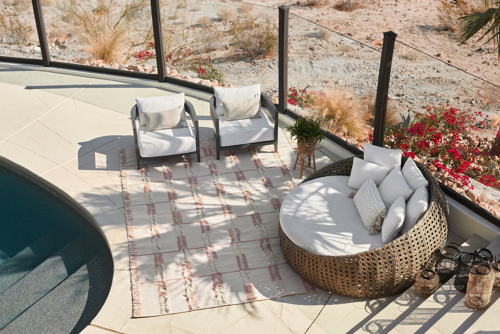 Round wicker outdoor daybed with white cushions and pillows, paired with two gray-framed chairs on a striped rug by a pool.