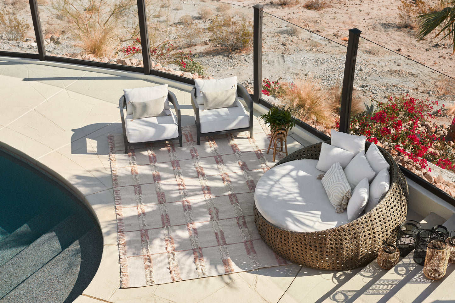 Poolside lounge with round brown wicker daybed and white cushions, two black-framed chairs, striped rug, potted plant.