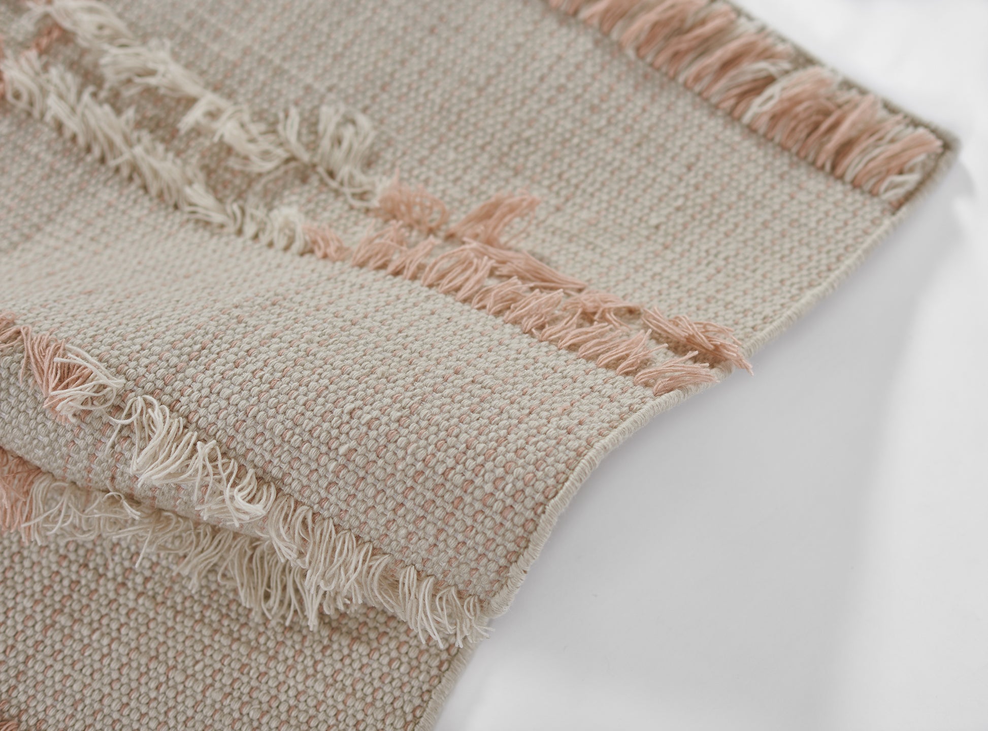 Soft beige woven rug with peach-pink fringe along edges and a textured, chunky weave.