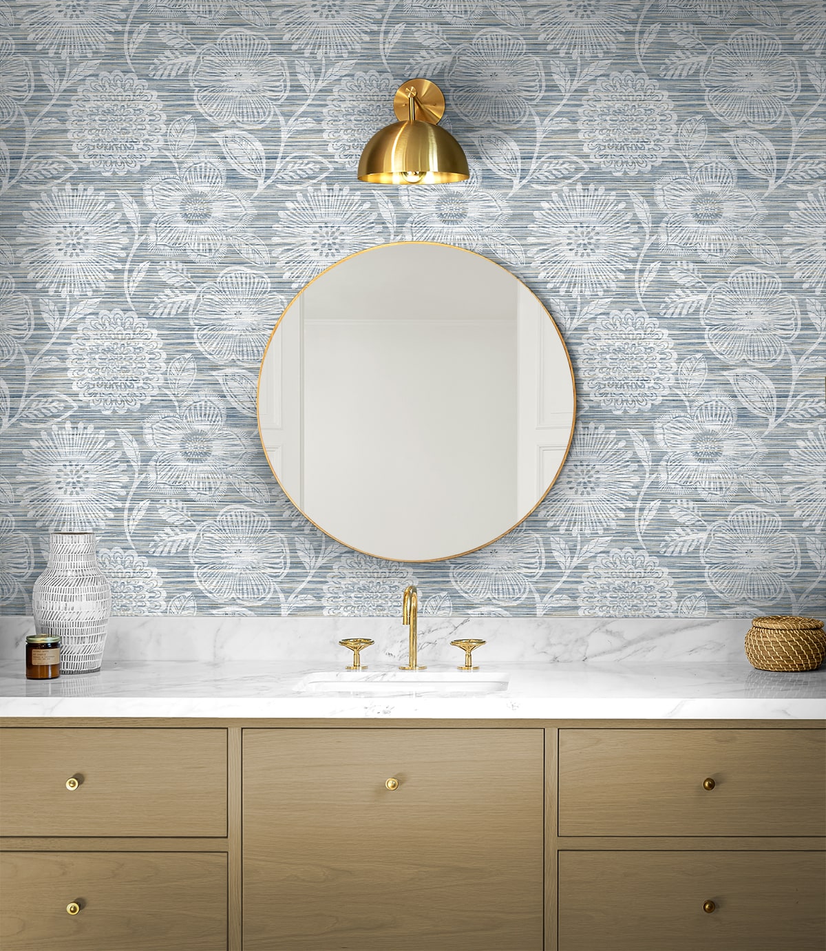 Round gold-framed mirror above marble countertop with gold faucet, blue floral wallpaper & light wood vanity.