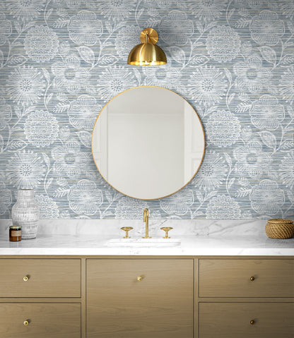 Round gold-framed mirror above marble countertop with gold faucet, blue floral wallpaper & light wood vanity.