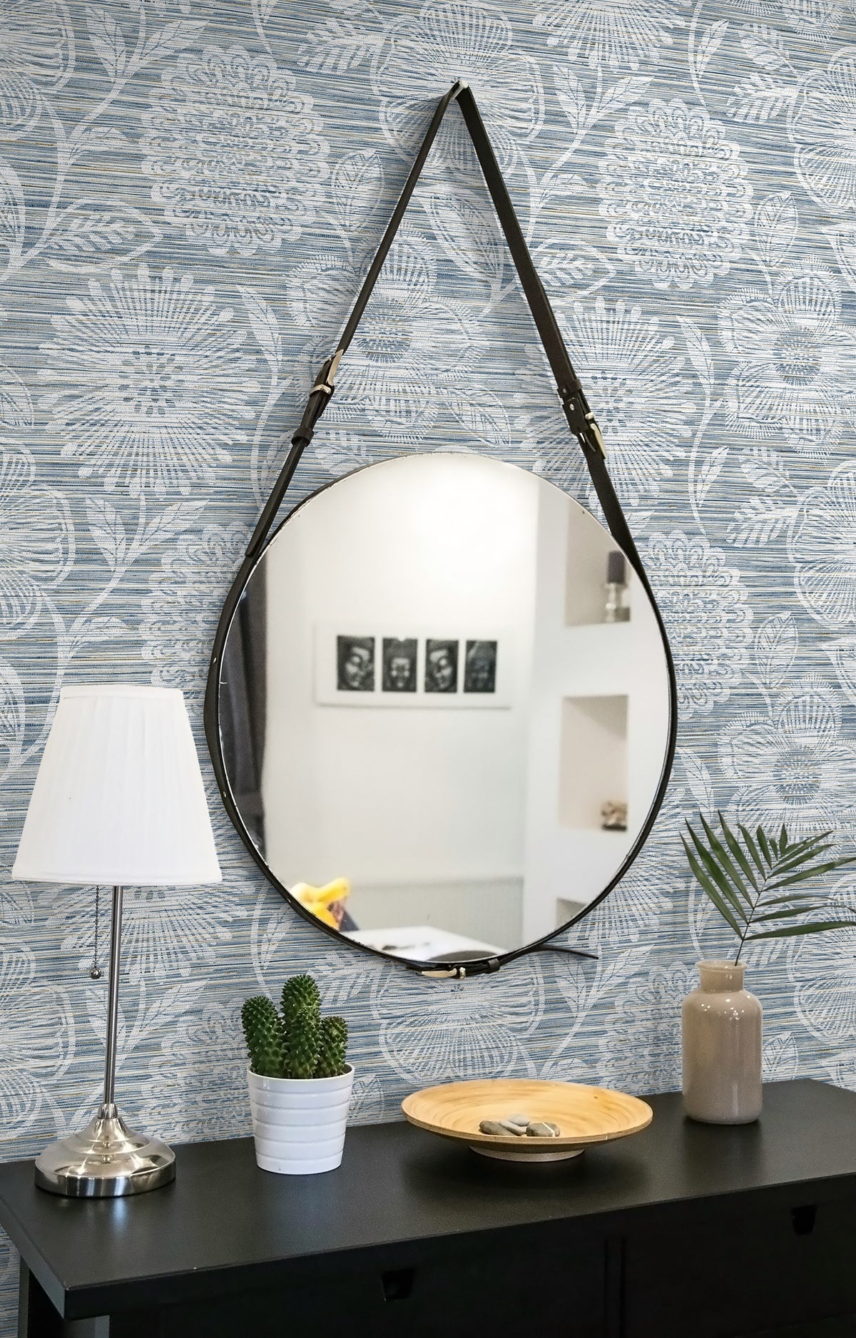 Blue floral wallpaper backdrop for a round mirror with a black metal frame and leather strap hung above a black console.
