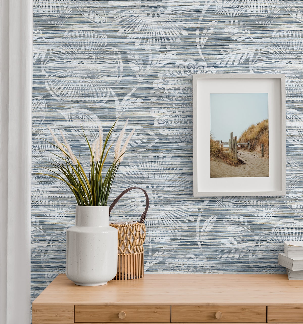 Coastal vignette with blue botanical wallpaper, light wood console, white ceramic vase, wicker basket, framed beach photo.