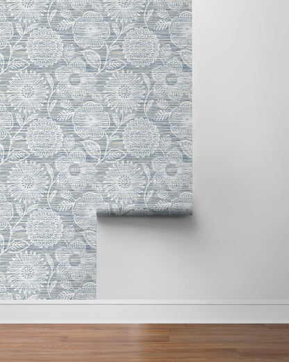 Blue-gray damask wallpaper roll with intricate medallion pattern, partially unrolled on a gray wall above a wood floor.