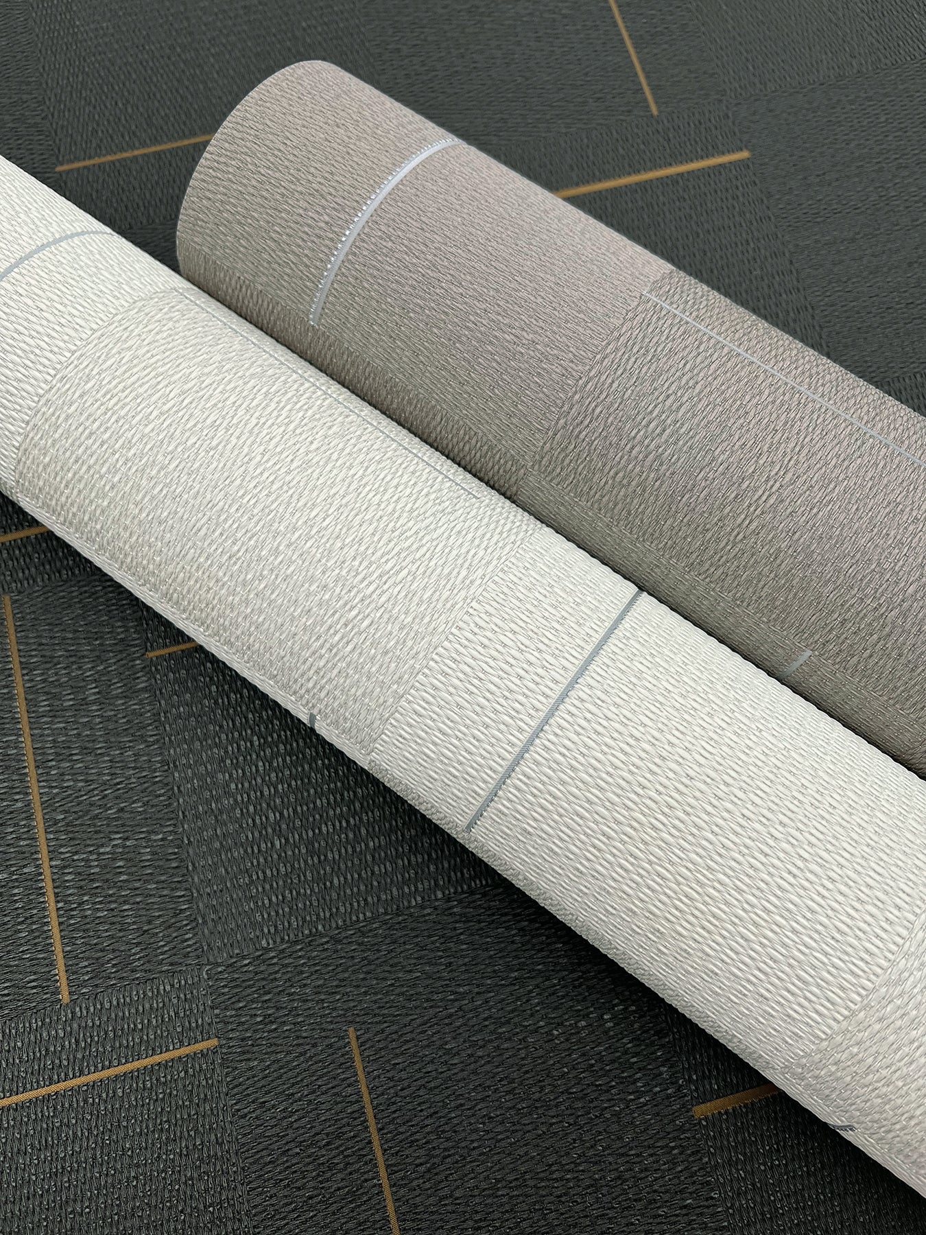 Two textured wallpaper rolls in ivory and taupe with a woven grain and visible seams on a dark surface.
