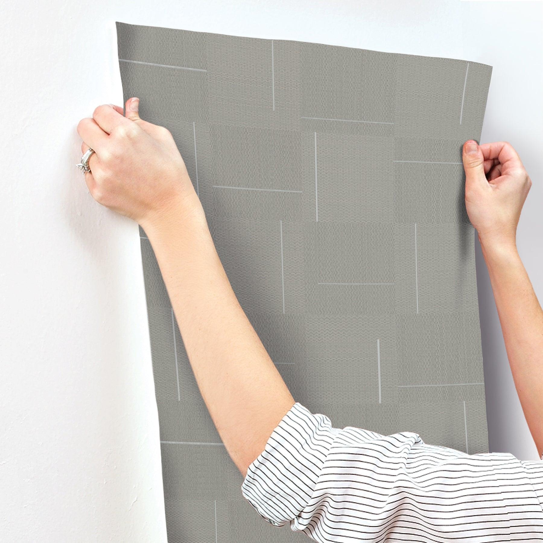 Two hands hold a large gray wallpaper sample with a geometric rectangle pattern against a white wall.