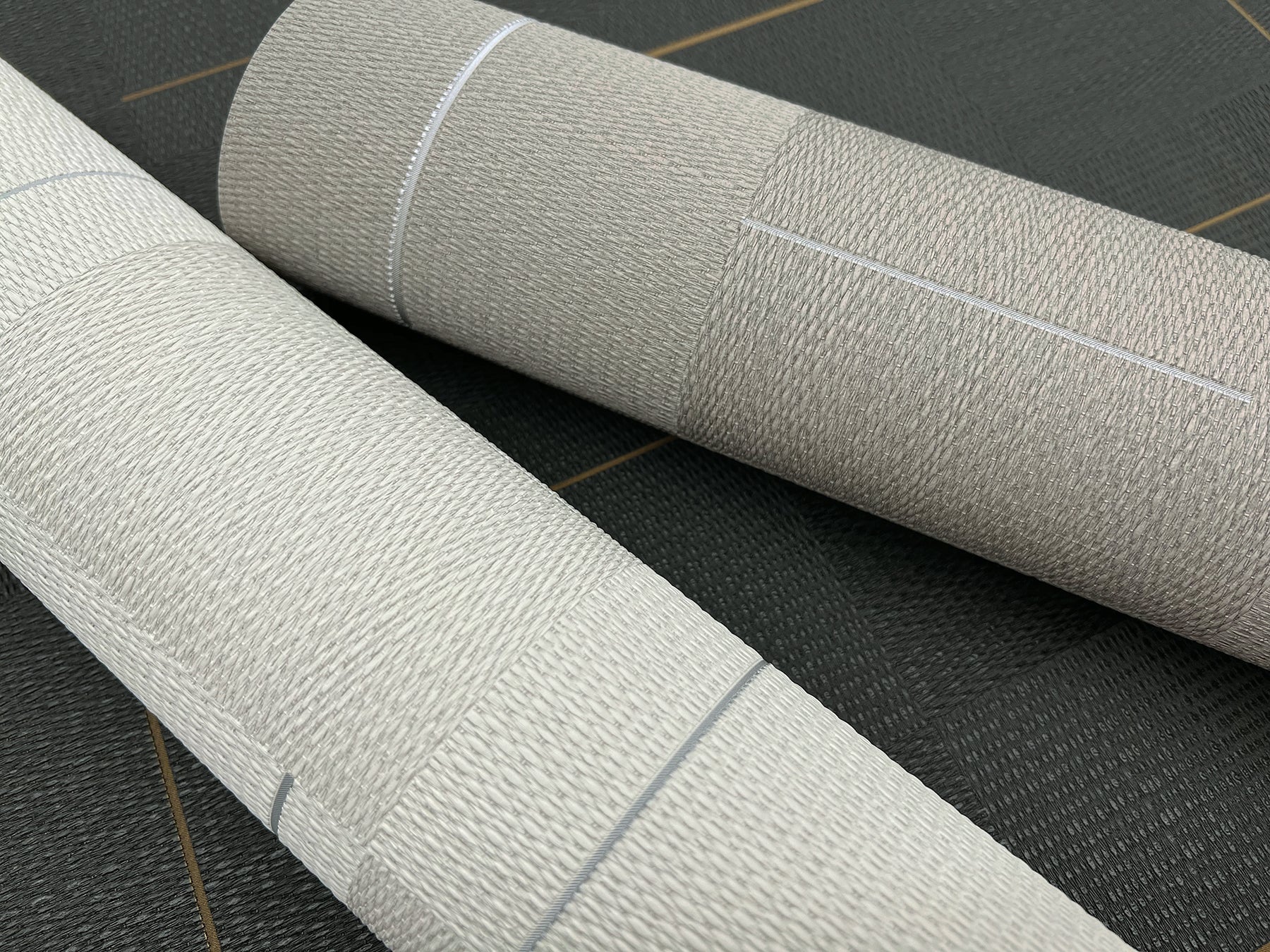 Two beige textured wallpaper rolls with woven linen pattern, lying on a dark surface.