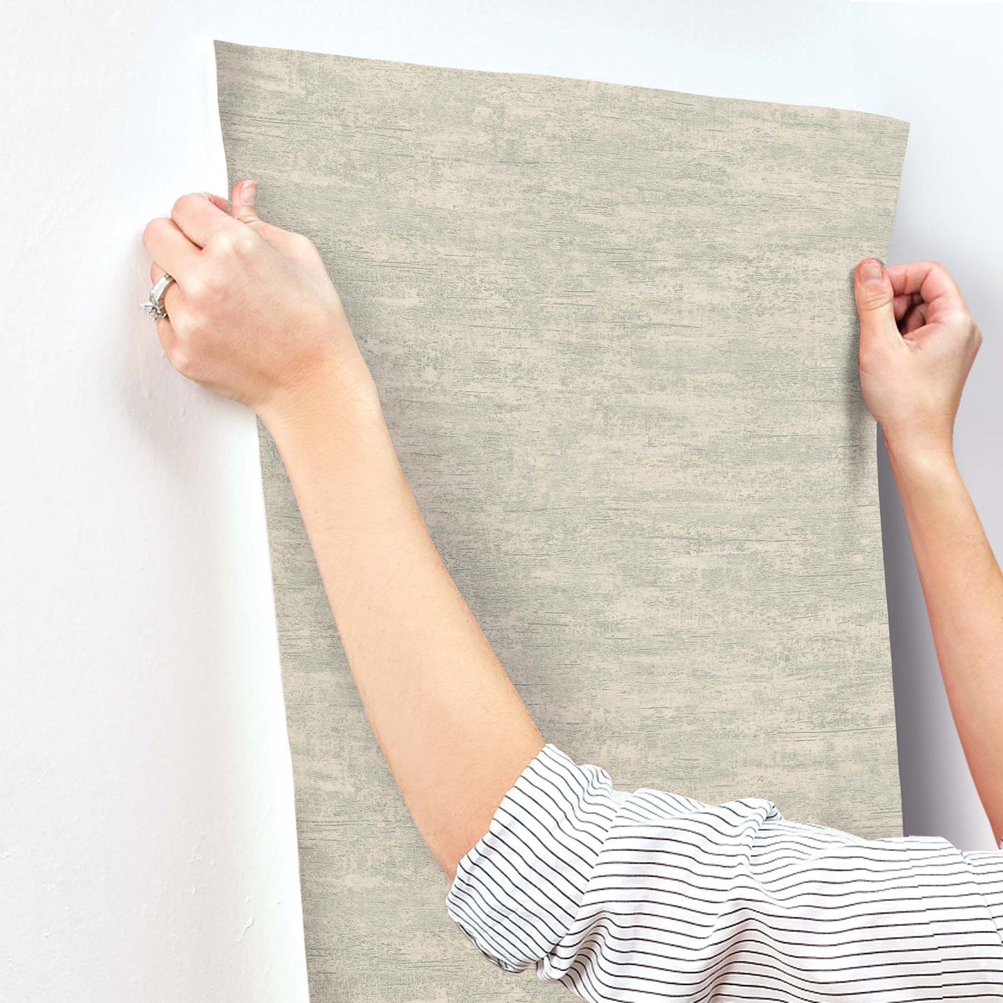Grey weathered stucco ecru wallpaper sample being held by hands with striped sleeves.