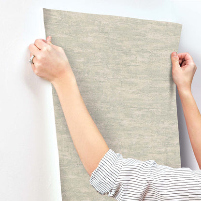 Grey weathered stucco ecru wallpaper sample being held by hands with striped sleeves.