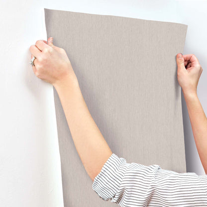 Light gray textured wallpaper with vertical ribs being applied to a wall; hands hold and position the large panel.
