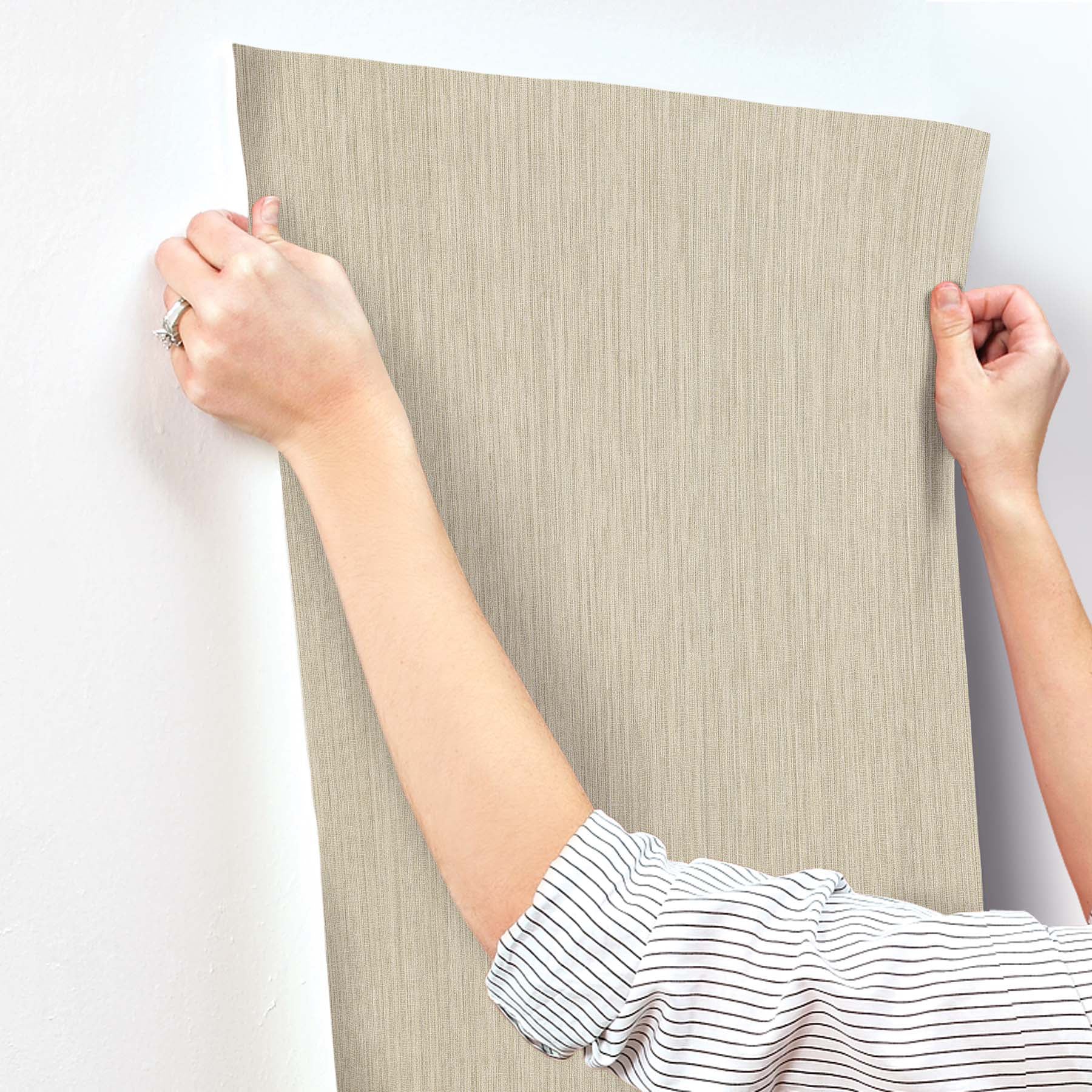 Beige linen-textured wallpaper panel being applied to a wall by two hands; striped sleeve visible.