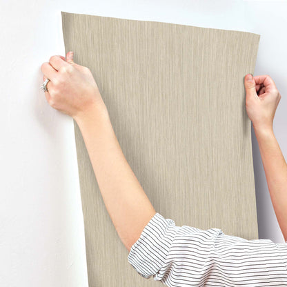 Beige linen-textured wallpaper panel being applied to a wall by two hands; striped sleeve visible.