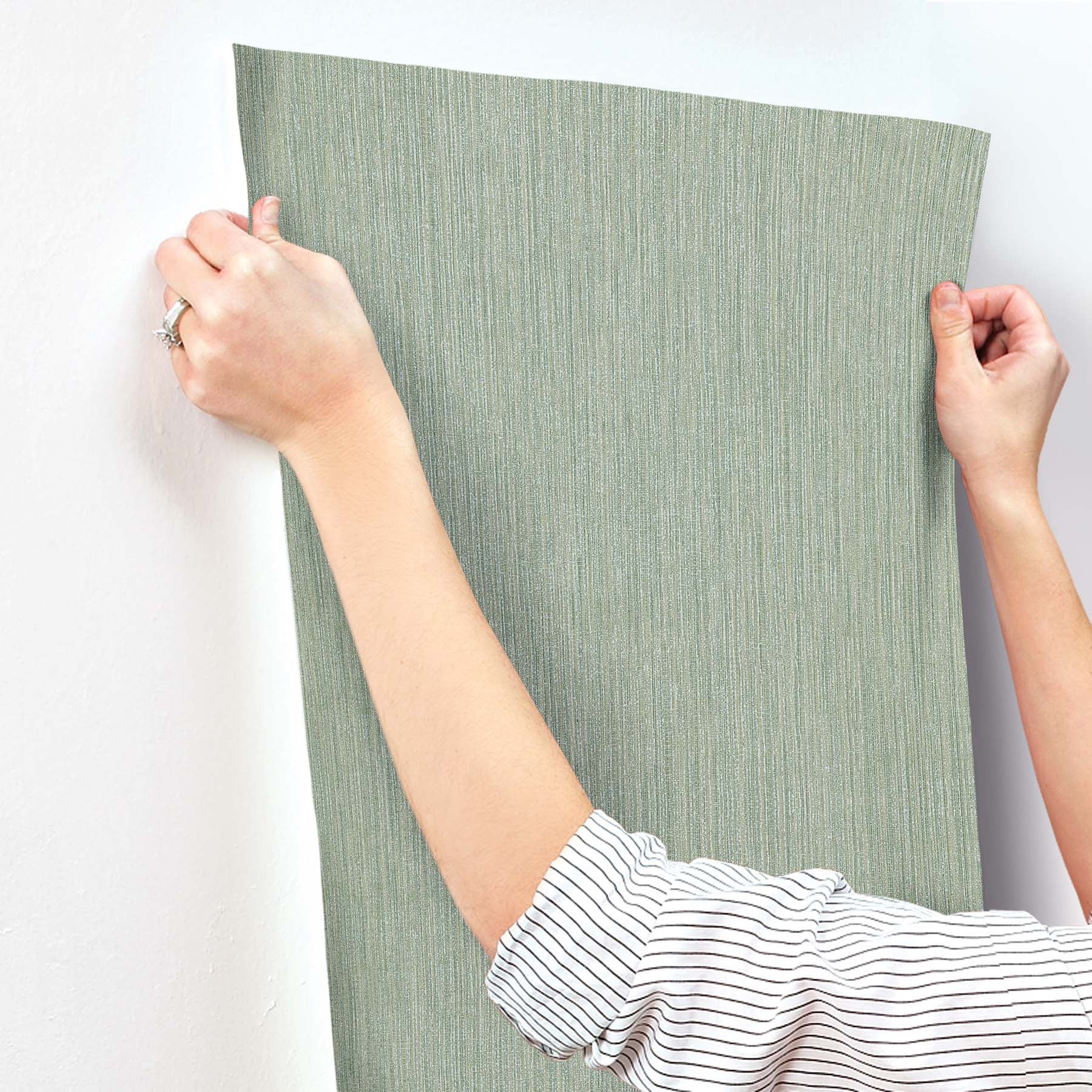 Green textured wallpaper with subtle vertical grasscloth grain, Sea Glass tones, held by a person wearing a striped shirt.