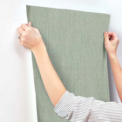 Green textured wallpaper with subtle vertical grasscloth grain, Sea Glass tones, held by a person wearing a striped shirt.
