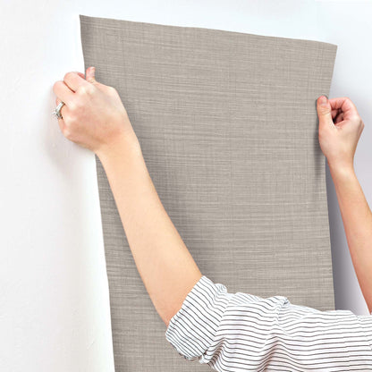 Beige linen-textured wallpaper sheet being applied to a wall; subtle crosshatched fabric look.
