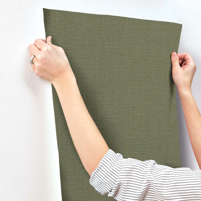 Olive-green textured wallpaper panel held by two hands, ready to stick to a wall.
