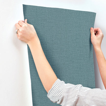 Teal textured wallpaper with a denim loom weave pattern, held against a white wall by hands wearing a striped shirt.