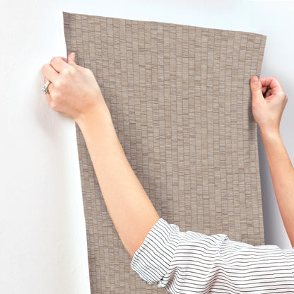 Hands holding a rectangular taupe wallpaper sample with a woven/grid texture; striped shirt sleeves visible.