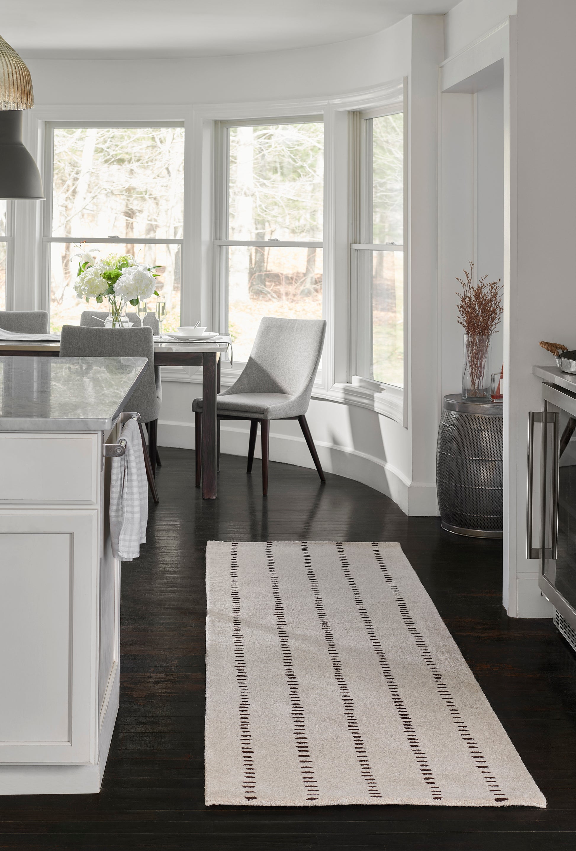 Cream woven area rug with dark brown dashed stripes on dark hardwood floor beside white kitchen island.