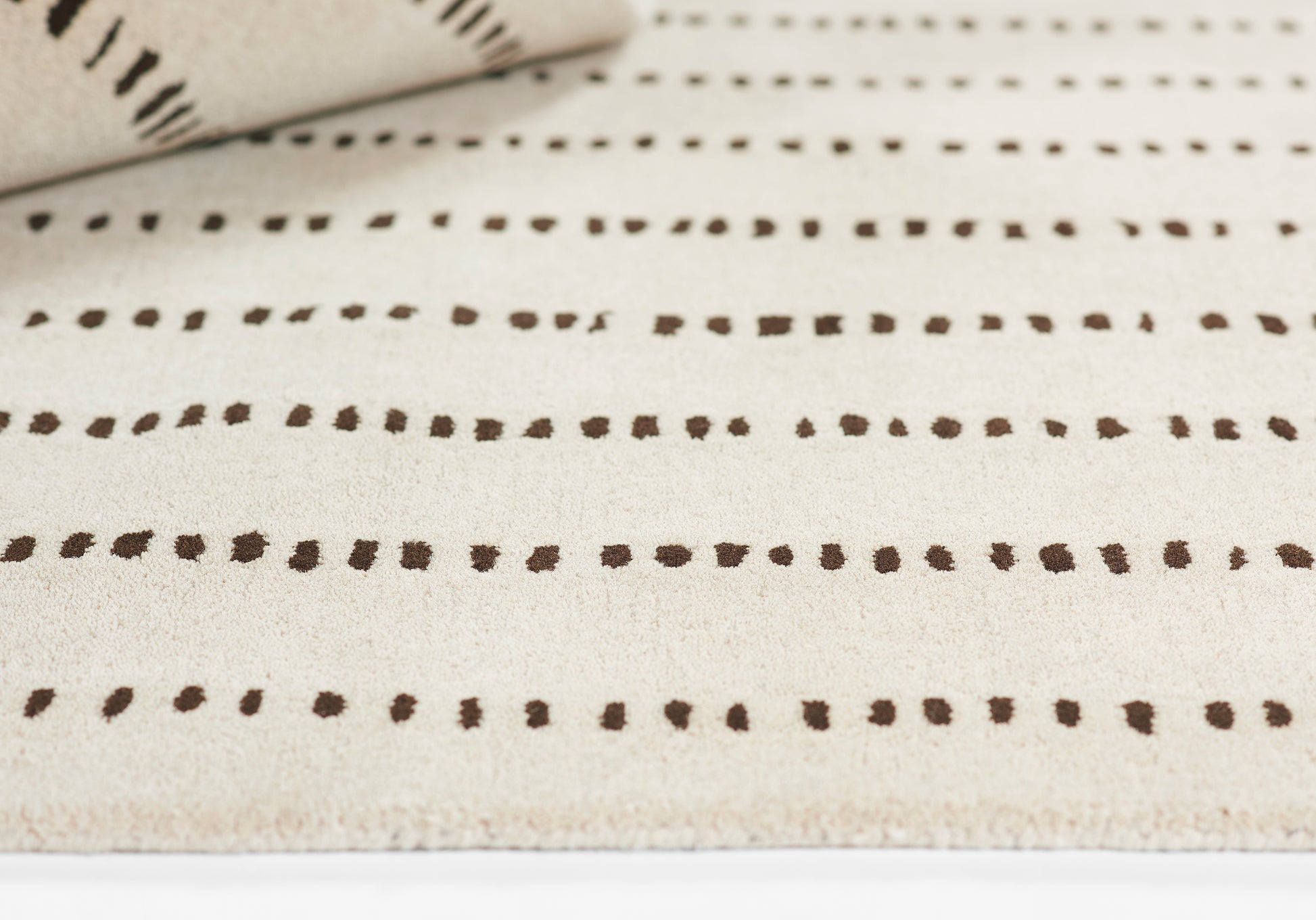 Beige rug with rows of dark brown dots on a light background; rolled corner visible at top-left.