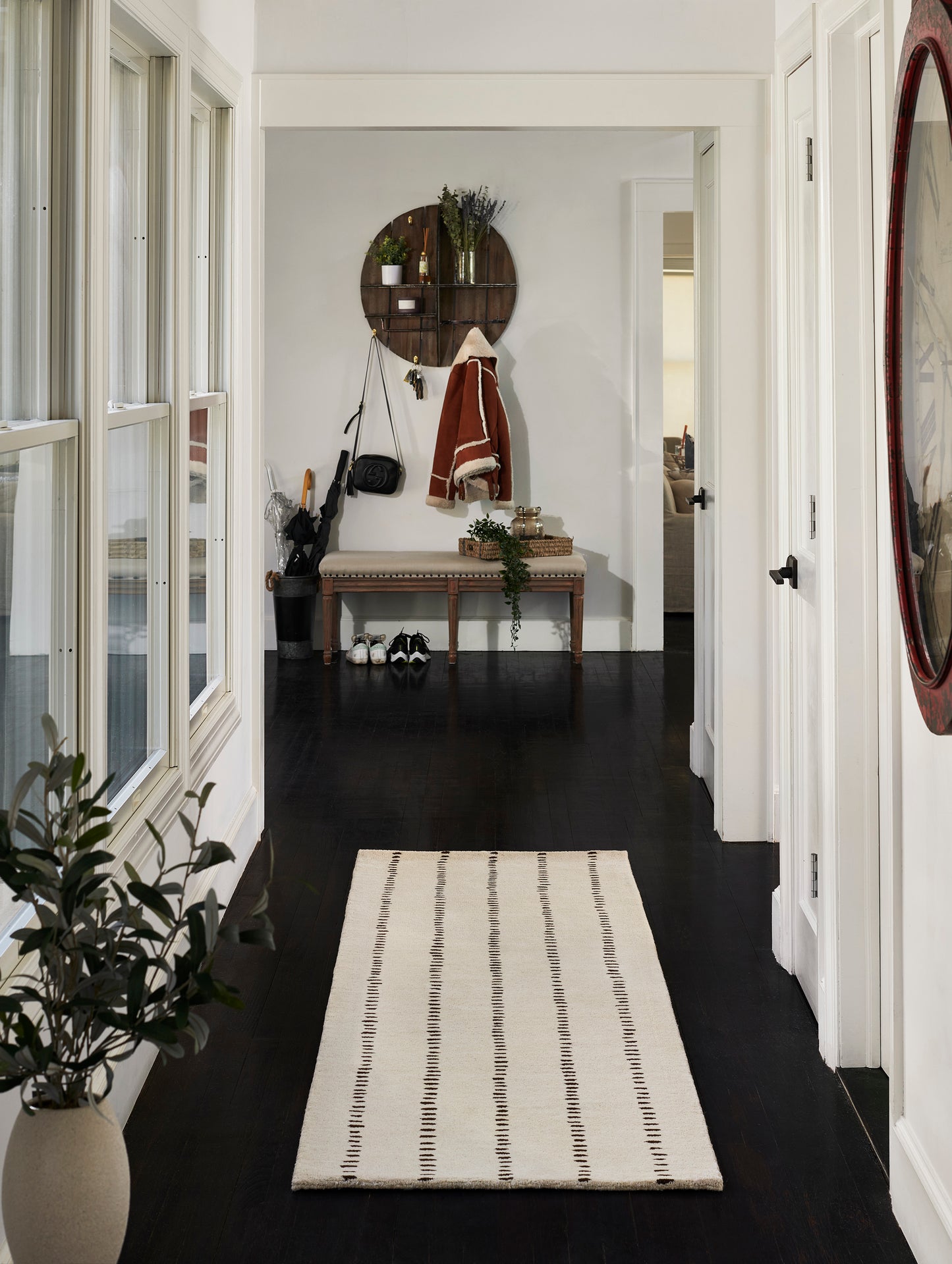 Cozy entryway with dark wood floor, cream striped runner, round wooden shelf, bench, coats and plants, red-framed mirror.