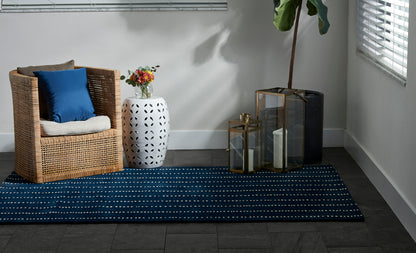 Woven wicker armchair with blue cushion, white perforated side table with flowers, navy rug, brass lanterns and tall plant by window.