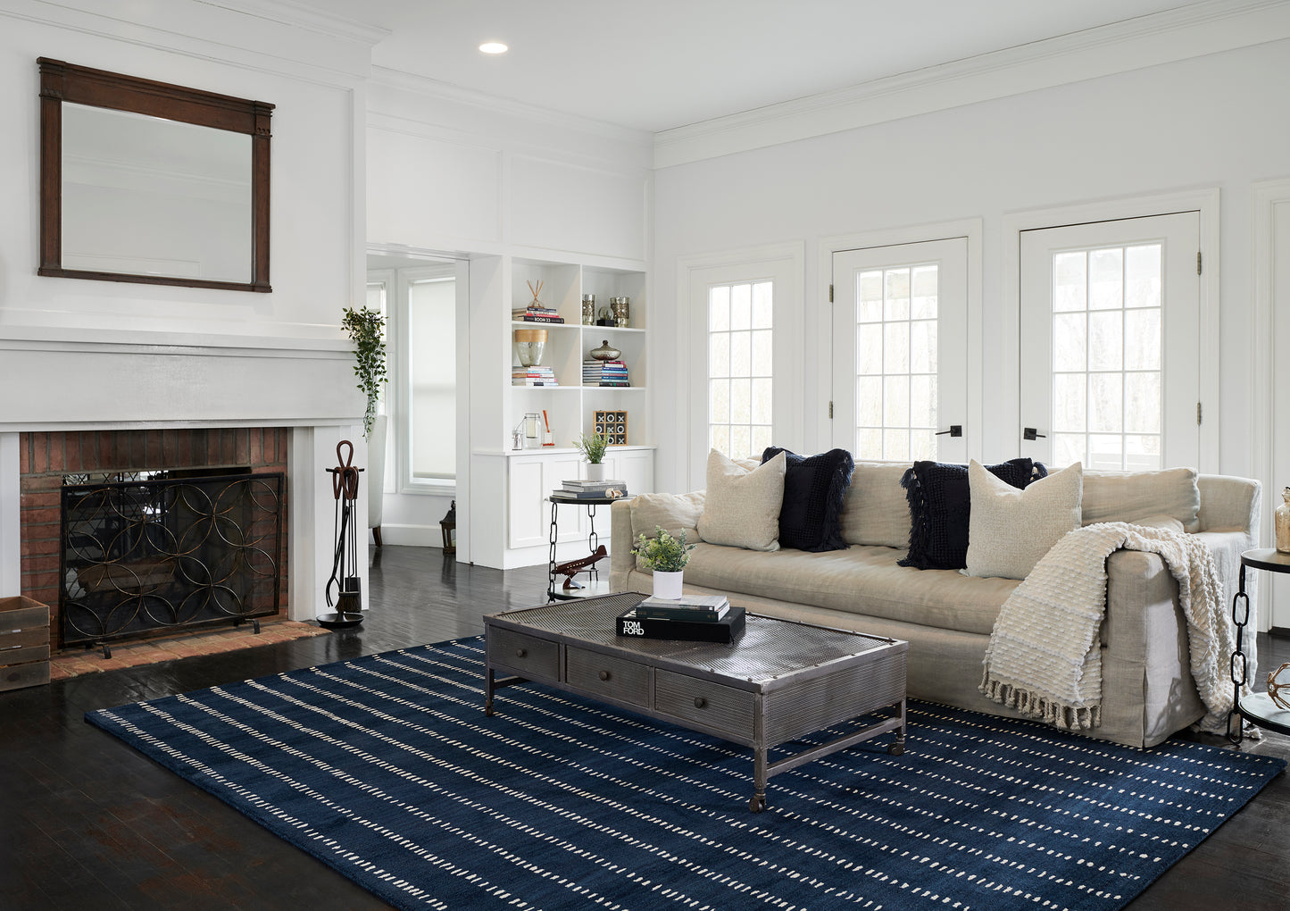 Bright living room with beige sofa, navy pillows, cream throw, dark metal-wood coffee table, blue striped rug, white walls.