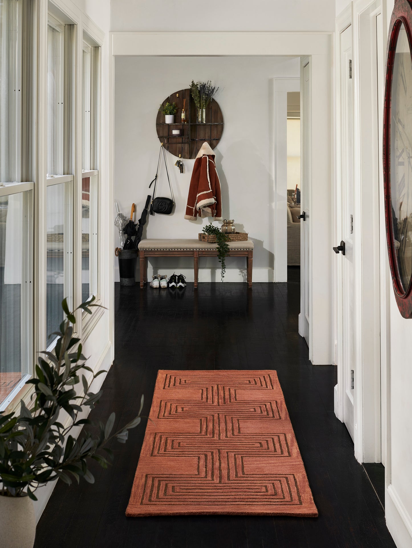 Orange geometric woven rug on dark wood hallway floor; entryway with wooden bench, round mirror, coat rack, and plants.