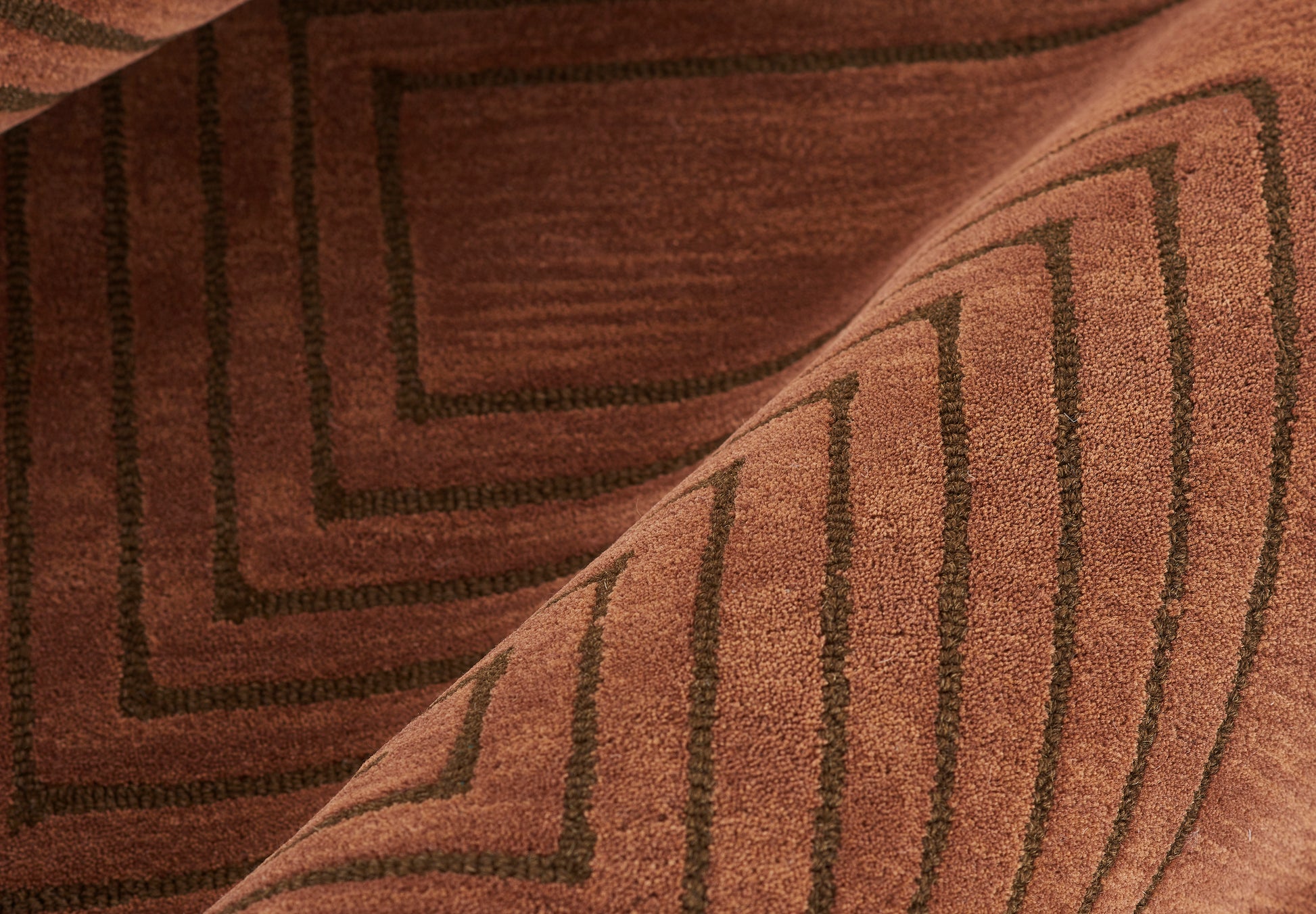 Brown woven rug with dark geometric rectangular border pattern, folded corner, warm earth tones.