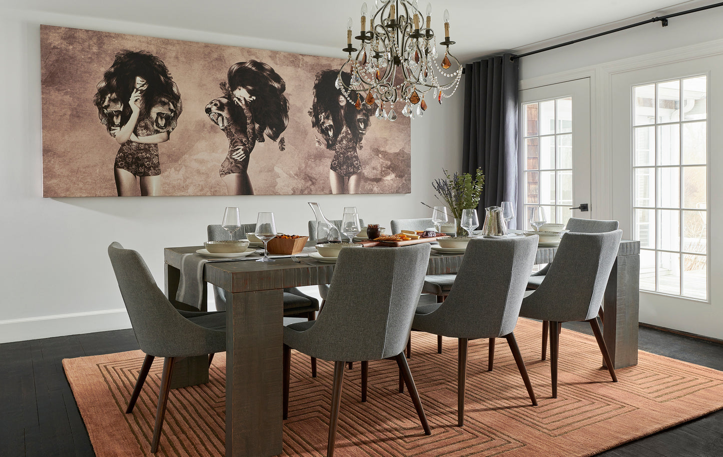 Long wooden dining table with gray upholstered chairs, a chandelier, orange rug, and a vintage mural of women on the wall