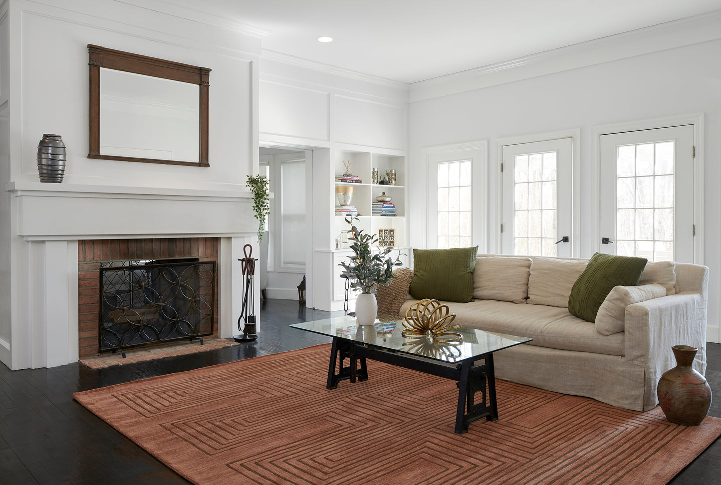Bright living room with white walls, brick fireplace, beige sofa, green pillows, glass coffee table, orange rug, built-ins.