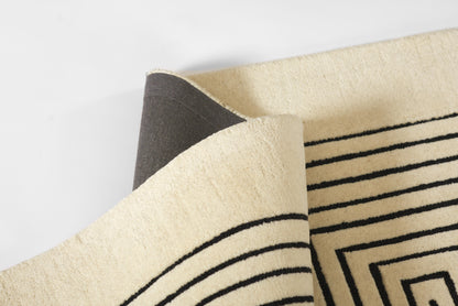 Beige rug with black geometric concentric square pattern and striped border, folded corner.