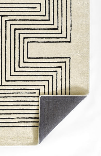 Rectangular beige rug with bold black geometric maze pattern; woven texture, folded corner reveals dark gray backing.