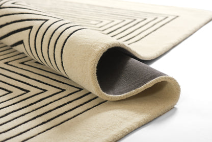 Beige rectangular rug with bold black geometric lines, rolled to reveal soft pile and dark underside; modern area rug.