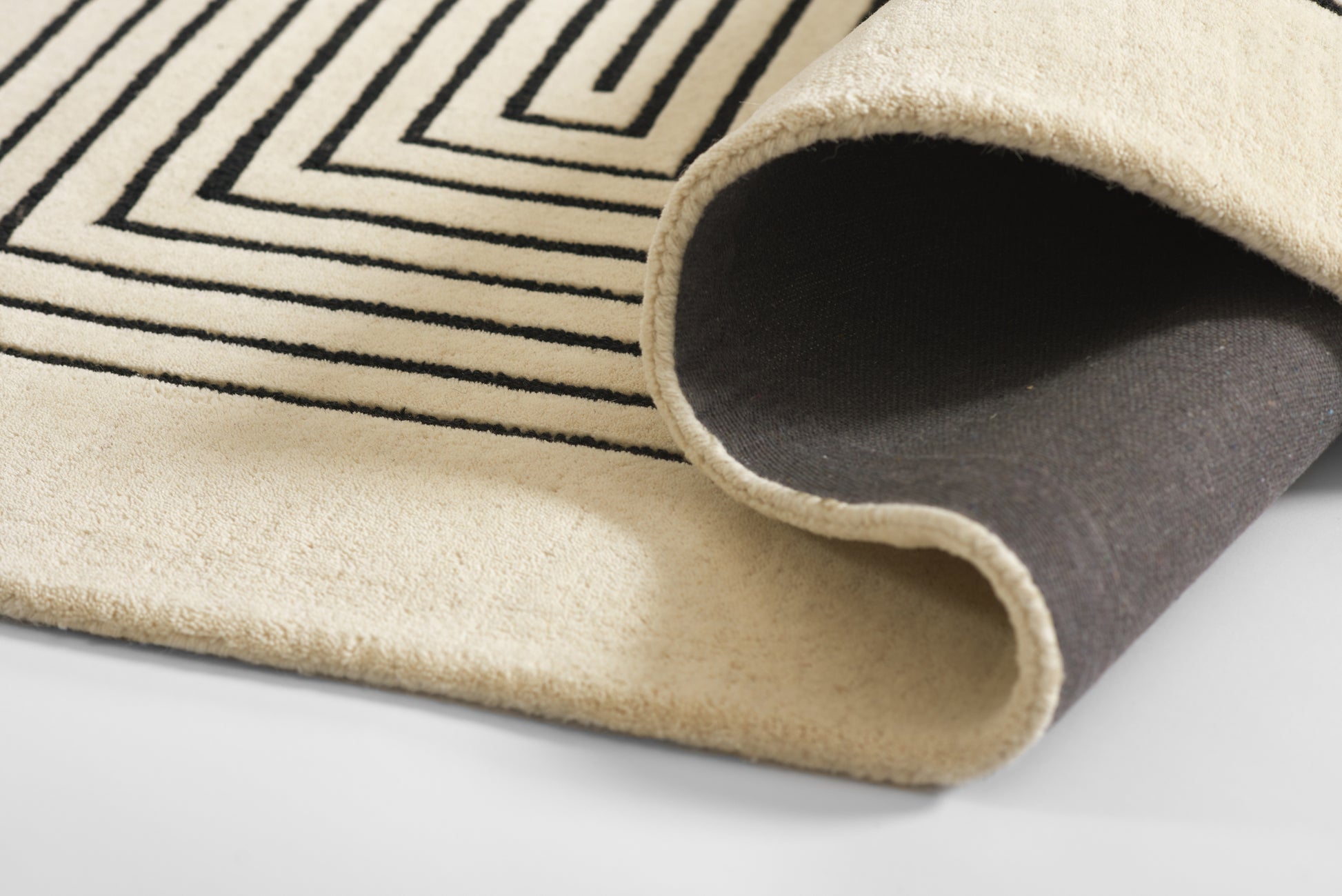 Beige wool-like area rug with black geometric linear pattern, rolled corner showing dark underside.