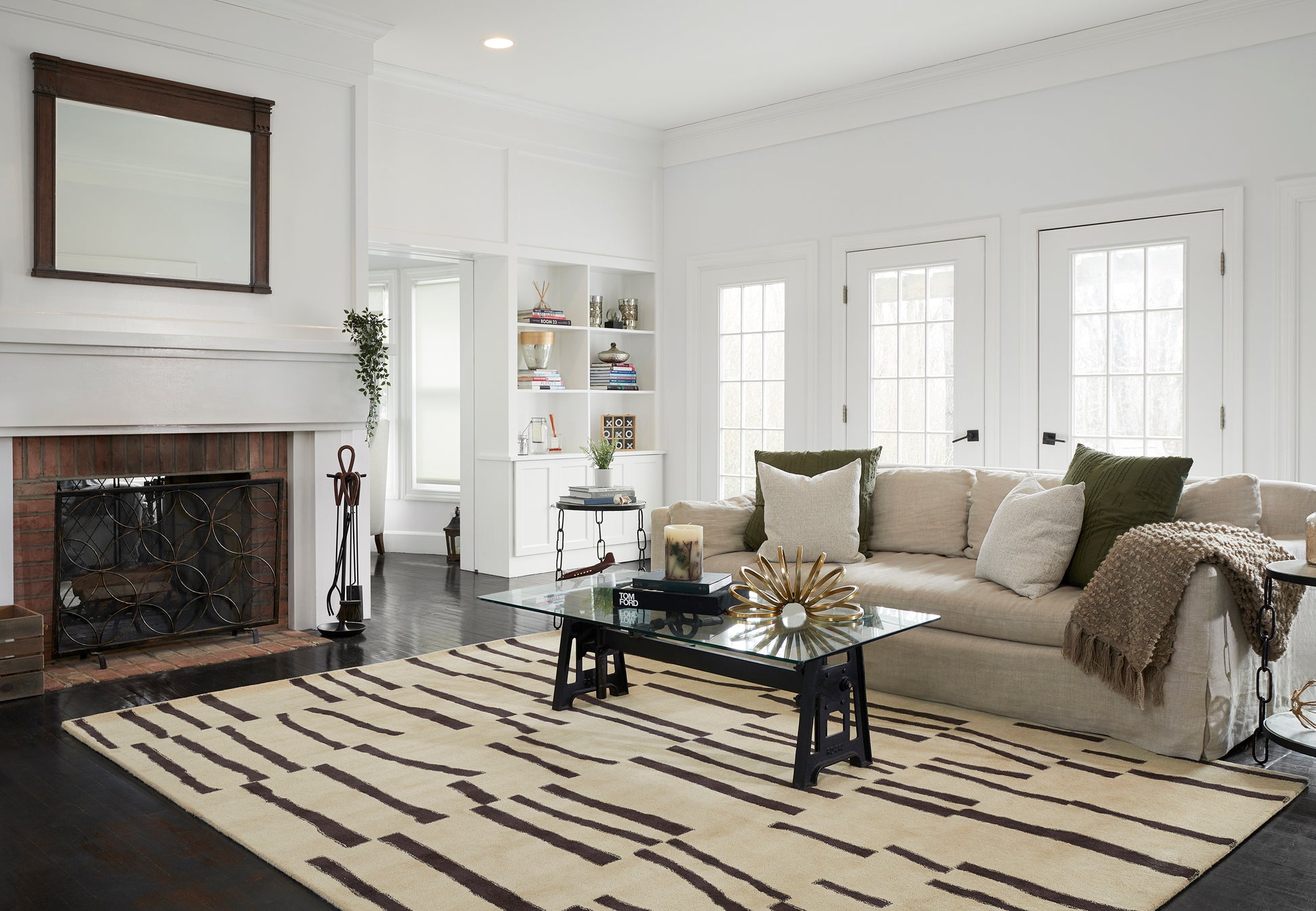 Bright living room with white walls, brick fireplace, dark wood mantel, beige sofa, glass coffee table, striped beige rug.