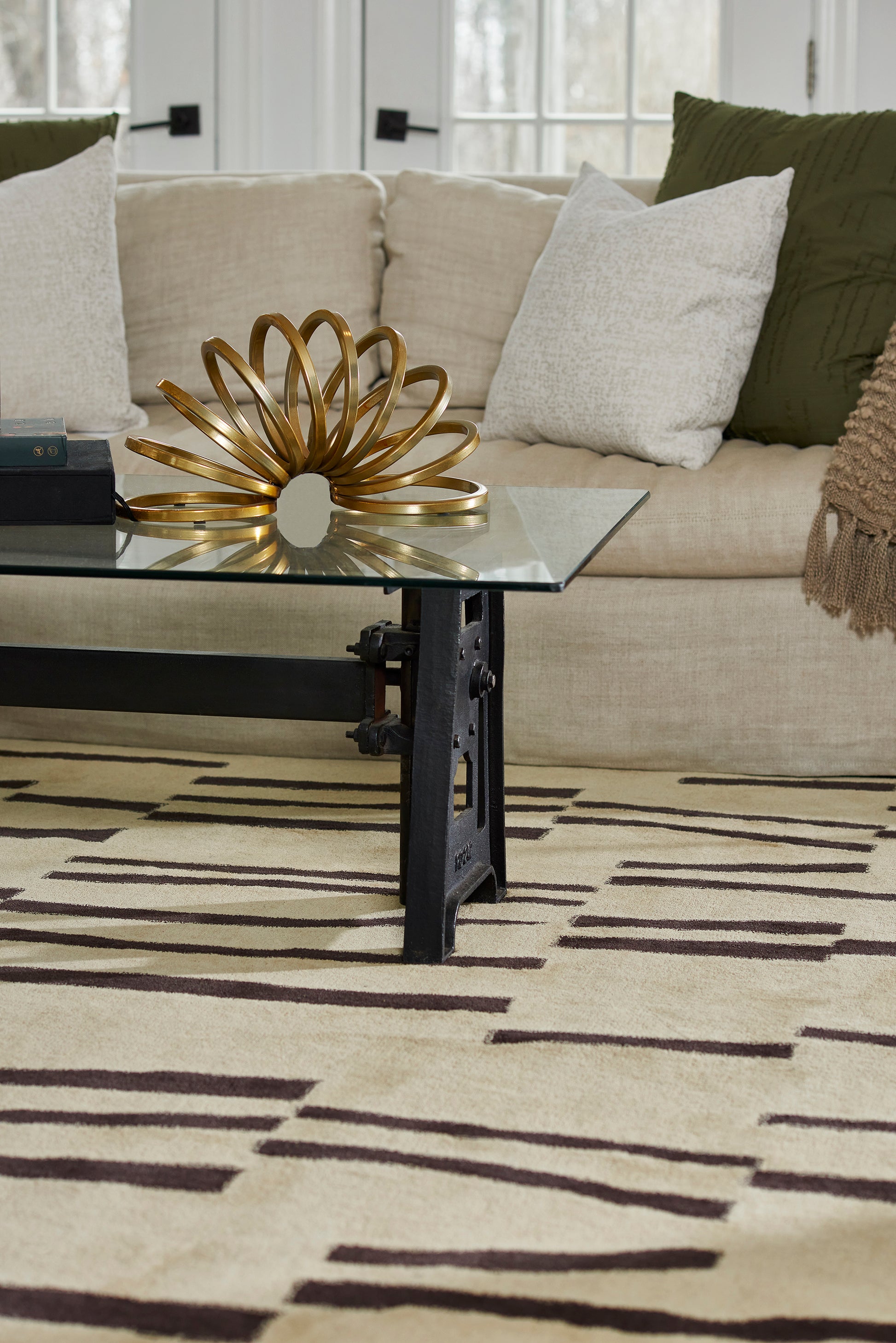 Gold looped metal sculpture on glass coffee table in a beige sofa living room with white & green cushions & a rug.