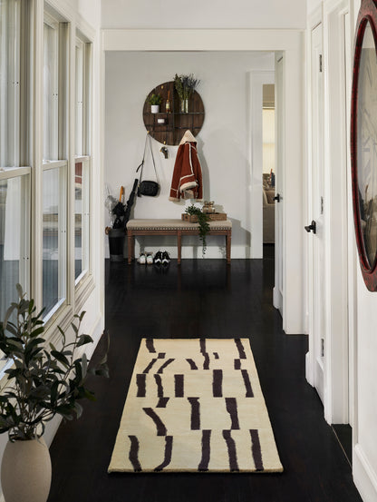 Hallway with dark wood floor, cream walls, beige runner rug with brown geometric lines, bench and round wall shelf.