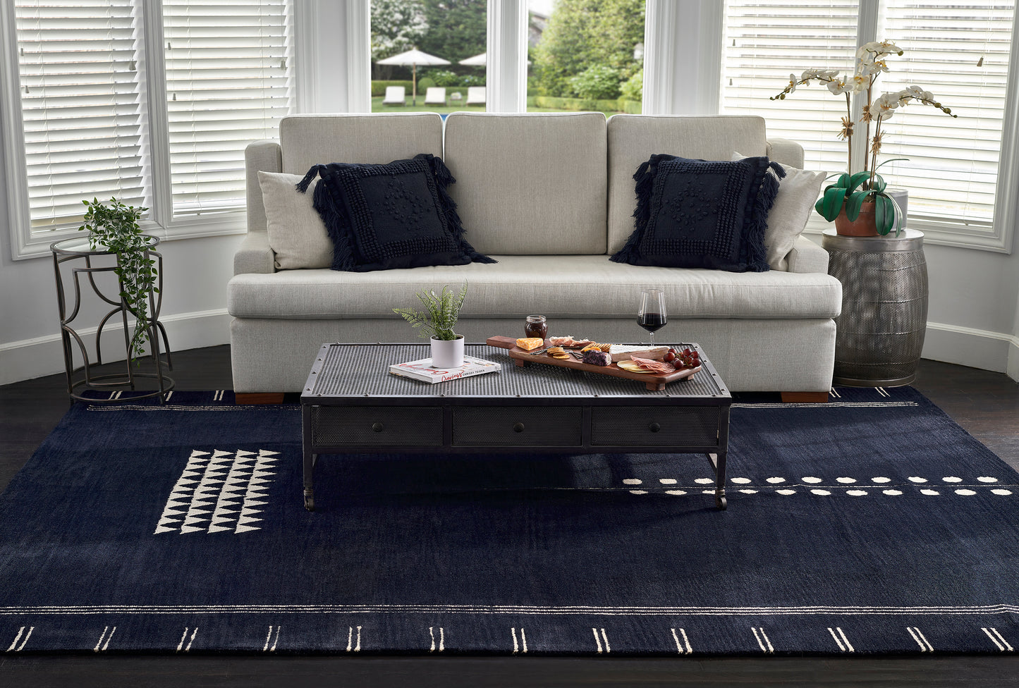 Gray fabric sofa with navy fringe pillows, black coffee table on a dark blue rug with geometric accents; plants and wine.