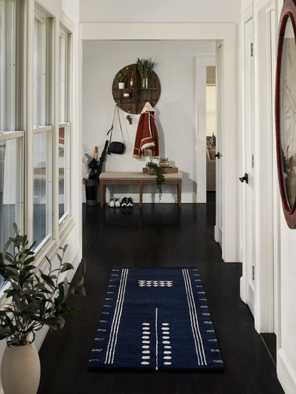Navy geometric area rug in a bright hallway with dark wood floor, bench, round wooden wall shelf and coat rack.