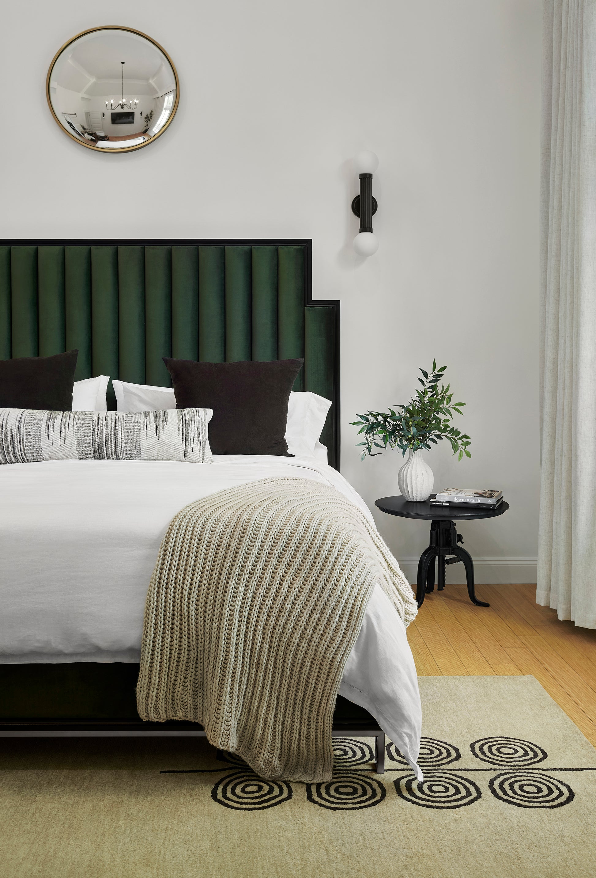 Bedroom with emerald green tufted headboard, white bedding, beige knit throw, dark pillows, gold mirror, black side table.