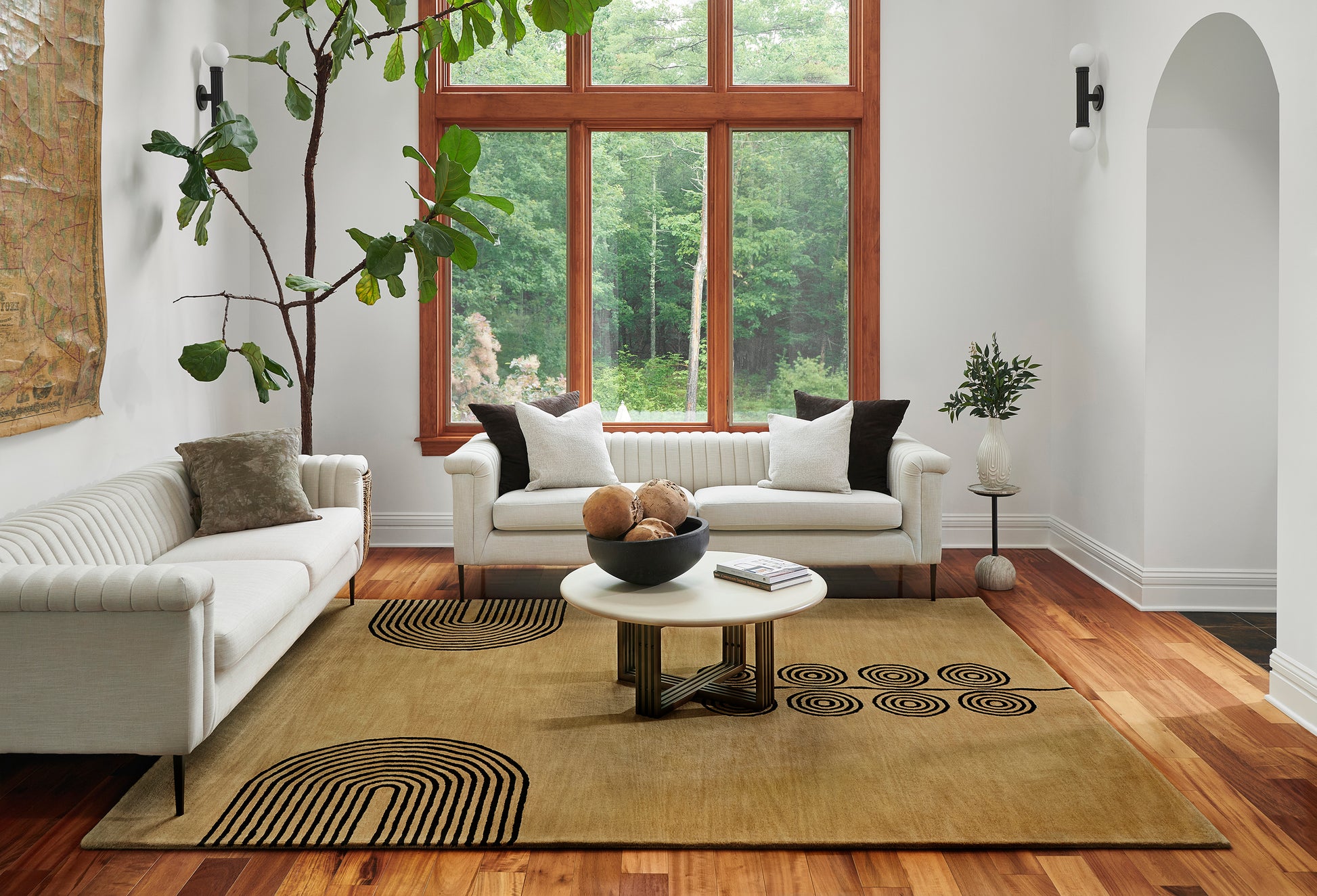 Bright living room with white sofas, wood-framed window, round white coffee table, tan rug with black arcs, potted plants.