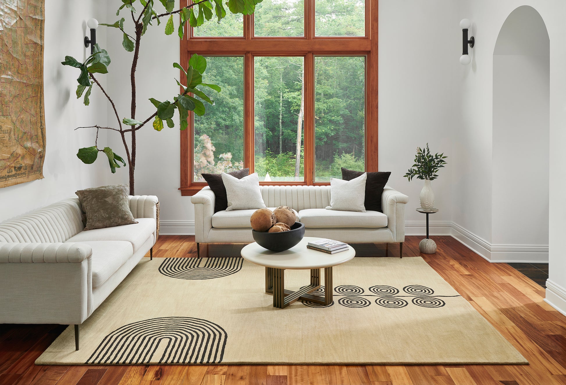 Bright modern living room with white tufted sofas, beige 5' x 8' rug, round coffee table, wooden floors, and a large wood-framed window.