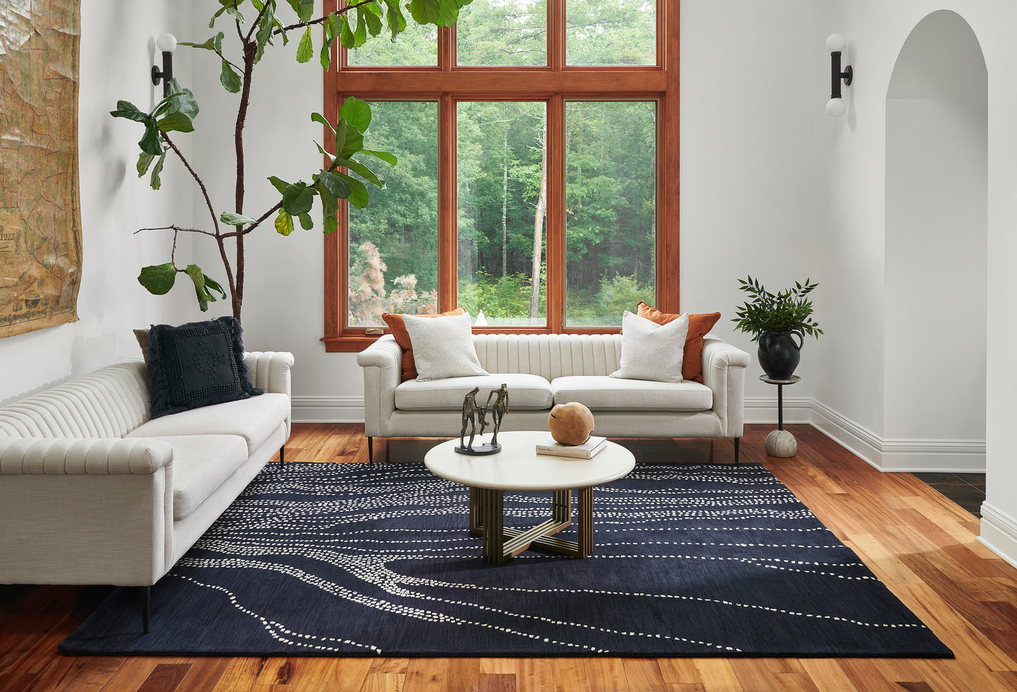 Bright living room with white sofas, navy runner rug, round coffee table, wood floors, large wood-framed windows & greenery.