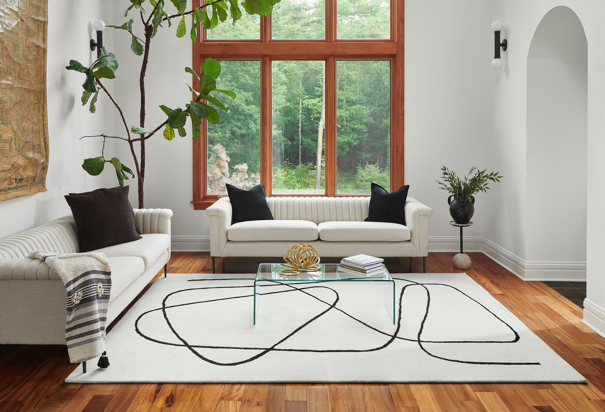 Bright modern living room with white sofas, glass coffee table, abstract black-lined rug, wood floor, and greenery.