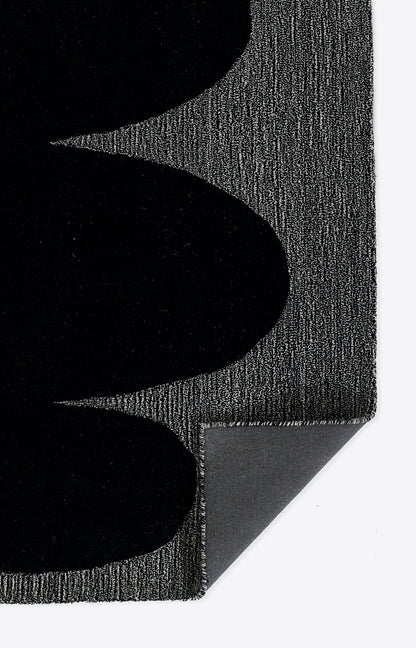 Close-up of a black textured rug with curved black shapes on a gray woven background; corner folded to reveal underside.