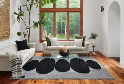 Bright living room with white sofas, glass coffee table, large gray rug with black oval patterns, wooden floors, and plants.
